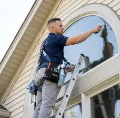 Professional window cleaner on a ladder using a squeegee to clean an exterior arched window, Jersey City, NJ.